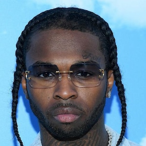 May include: A close-up portrait of a person with black hair styled in braids, wearing gold-rimmed sunglasses with a dark lens. The person has a dark complexion and a beard.
