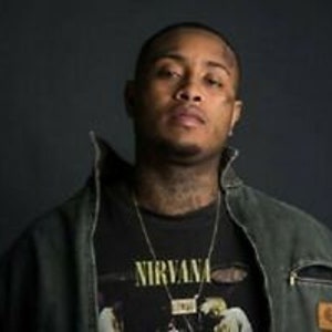 Puede incluir: Un hombre de tez oscura y cabello negro corto lleva una camiseta negra con la palabra "NIRVANA" impresa. También lleva una chaqueta verde oscuro y un collar de cadena plateada.