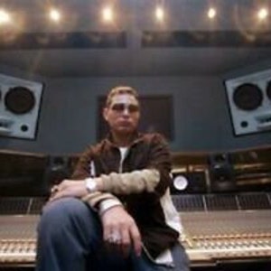 May include: A man sitting in a recording studio with a large mixing board in front of him. He is wearing a brown jacket and jeans. There are two large speakers on either side of him.