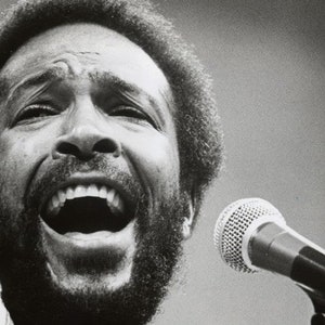 Può includere: Una fotografia in bianco e nero di un uomo che canta in un microfono. Indossa una camicia bianca e ha la barba e i capelli afro.