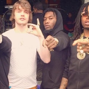 May include: Four men are standing together, looking at the camera. The man on the left is wearing a black jacket with a white and black patterned shirt underneath. He is wearing sunglasses and has a gold chain around his neck. The man in the middle is wearing a white t-shirt and has his hand raised in a gesture. The man on the right is wearing a black jacket with a gold chain around his neck. He is wearing sunglasses and has dreadlocks. The man in the back is wearing a black jacket and has his hand raised in a gesture.