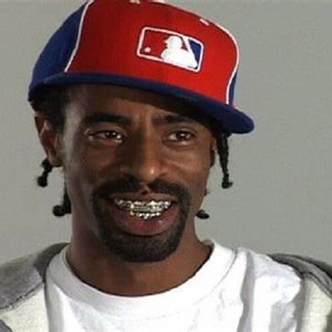 Könnte beinhalten: Ein Mann mit einem Ziegenbart und einer Baseballkappe mit einem rot-weiß-blauen Logo lächelt. Er trägt ein weißes T-Shirt und einen grauen Hoodie. Er hat einen silbernen Grill auf seinen Zähnen.