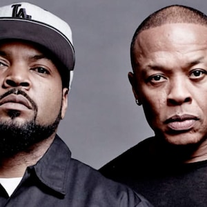 May include: A close-up portrait of two men, one wearing a black baseball cap with a white LA logo and a black shirt, the other wearing a black t-shirt.