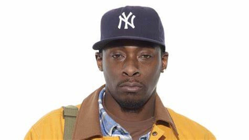 May include: A man wearing a navy blue baseball cap with a white "NY" logo. He is wearing a yellow jacket over a plaid shirt.