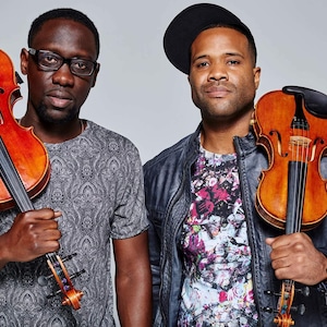 May include: Two men, one wearing a black baseball cap and a black leather jacket, and the other wearing a gray patterned shirt, are holding violins. Both violins are brown with black strings.