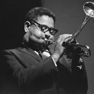 Könnte beinhalten: Ein Schwarzweißfoto eines Mannes, der Trompete spielt. Er trägt einen Anzug und eine Brille. Die Trompete wird in seiner rechten Hand gehalten und der Schalltrichter zeigt nach rechts.