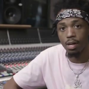 May include: A young man with a black bandana and a silver chain necklace is wearing a pink t-shirt. He is sitting in front of a soundboard in a recording studio.