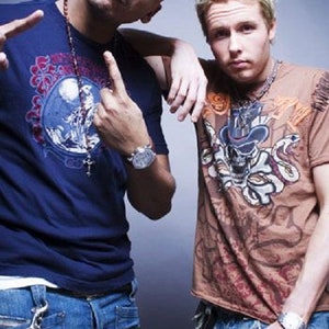 May include: Two men wearing casual clothing. The man on the left is wearing a blue t-shirt with a graphic print, a fedora hat, and blue jeans. The man on the right is wearing a brown t-shirt with a skull graphic, and blue jeans.