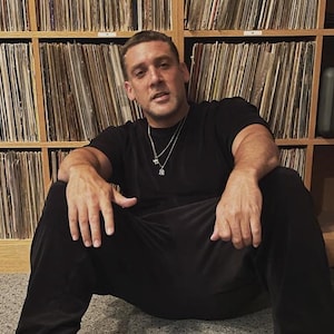 Puede incluir: Un hombre con una camiseta negra y pantalones negros se sienta frente a una pared de discos de vinilo. Lleva una cadena de plata con un pequeño colgante.