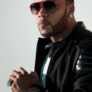 May include: A man wearing a black leather jacket with a white t-shirt underneath. He is wearing red sunglasses and a diamond earring. He has a short beard and is looking at the camera.