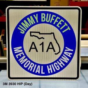 May include: A white square sign with a blue circle containing the text "JIMMY BUFFETT MEMORIAL HIGHWAY" in white and green lettering. Inside the circle is a black outline of the state of Florida with the text "A1A" in black.