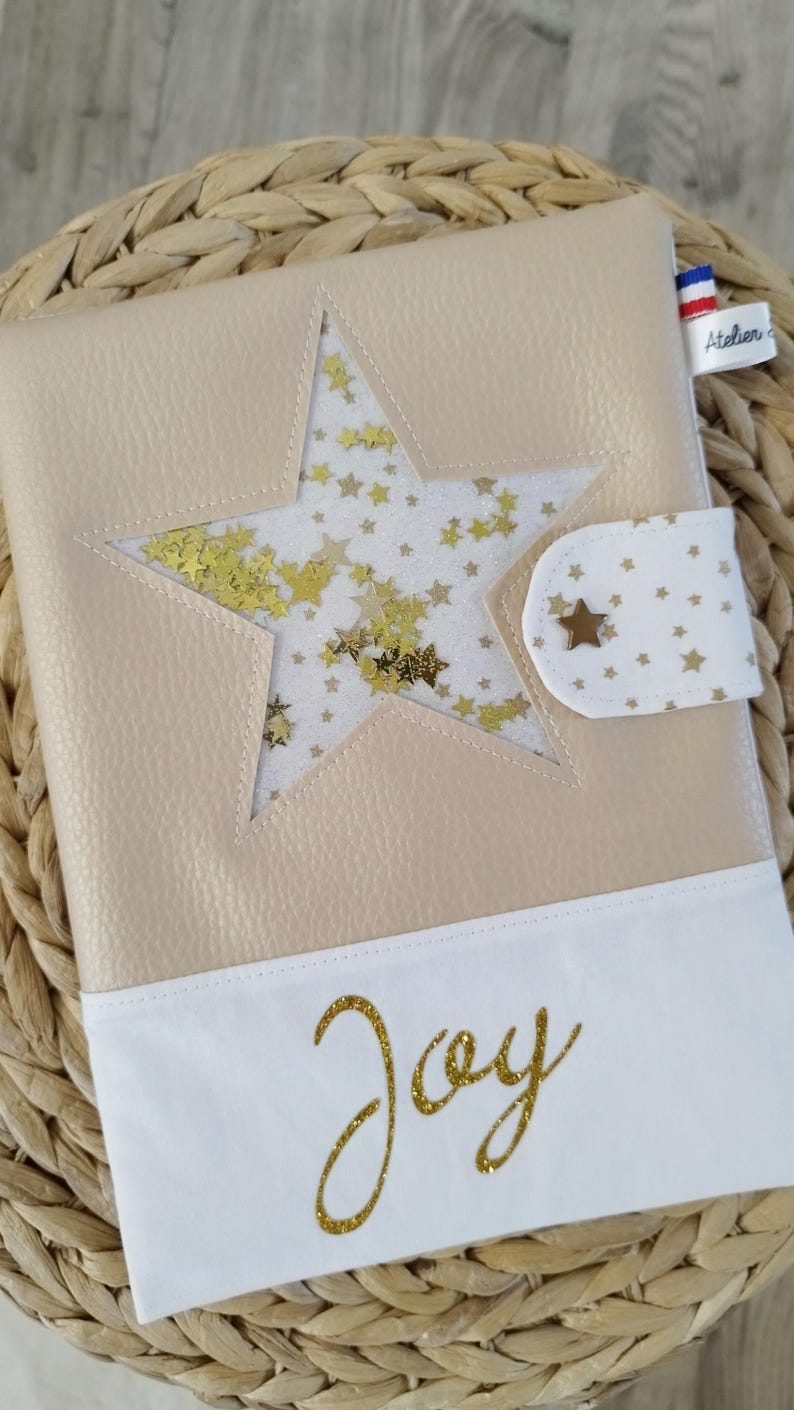 May include: A beige leatherette notebook with a white fabric flap. The flap has a gold glitter star design and the word "Joy" in gold glitter. The notebook has a clear window showing gold glitter stars on a white background.