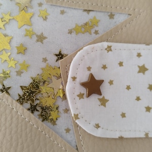 May include: A beige leather notebook with a white fabric flap featuring a gold star and a white background with gold stars. The flap is attached to the notebook with a gold star button.