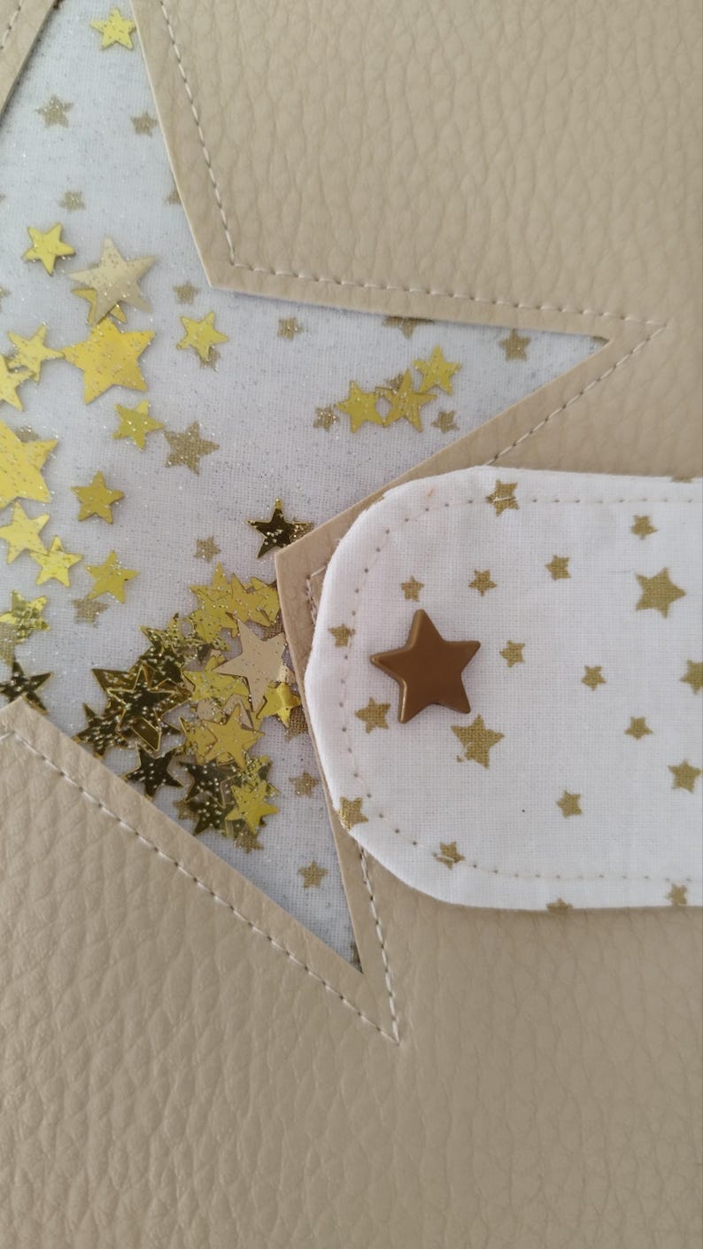May include: A beige leather notebook with a white fabric flap featuring a gold star and a white background with gold stars. The flap is attached to the notebook with a gold star button.