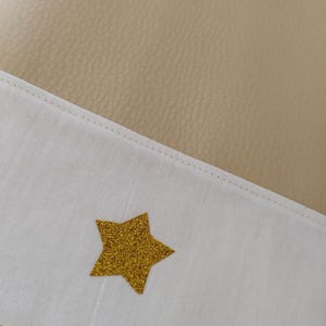 May include: A beige faux leather journal with a white fabric flap. The flap has a gold glitter star sewn on it.
