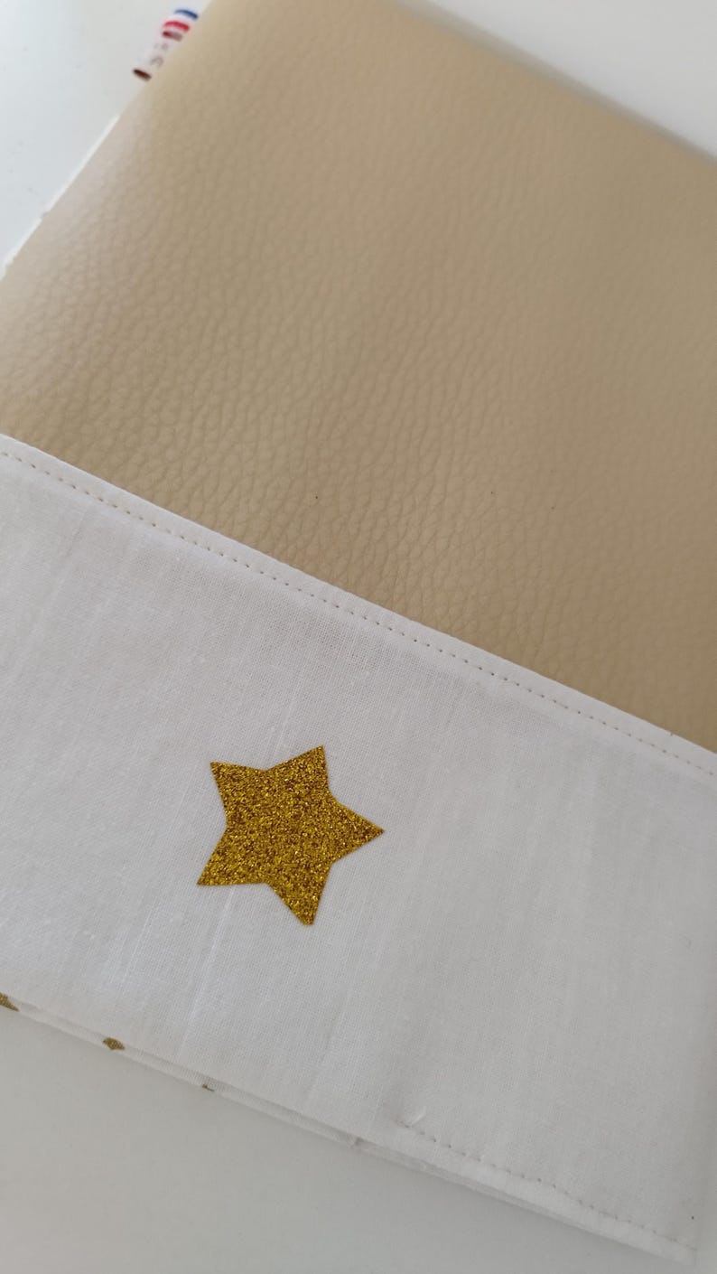 May include: A beige faux leather journal with a white fabric flap. The flap has a gold glitter star sewn on it.