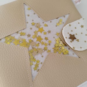 May include: A beige leather notebook with a star-shaped window revealing gold glitter stars on a white background. The notebook has a white fabric flap with a gold star button and the text "Atelier Scrap" in a white rectangle with a French flag.