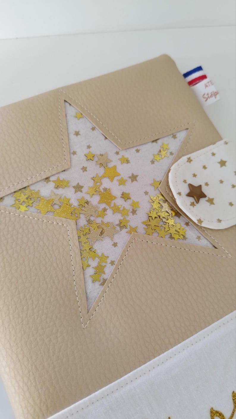 May include: A beige leather notebook with a star-shaped window revealing gold glitter stars on a white background. The notebook has a white fabric flap with a gold star button and the text "Atelier Scrap" in a white rectangle with a French flag.