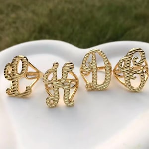May include: Four gold-colored initial rings, each featuring a different letter: L, H, D, and G. The rings are displayed on a white surface, with a blurred green background. The letters have a textured, striped design.