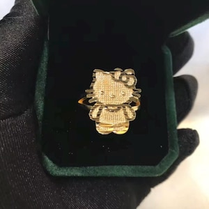 May include: A gold-colored ring featuring a Hello Kitty design. The ring is set in a dark green velvet box, with the ring's design clearly visible. The ring has a textured surface and is presented against a dark background.