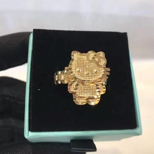 May include: A gold-colored ring featuring a Hello Kitty design. The ring is in a box with a black interior and a light blue exterior. The ring's design includes a bow and textured details.