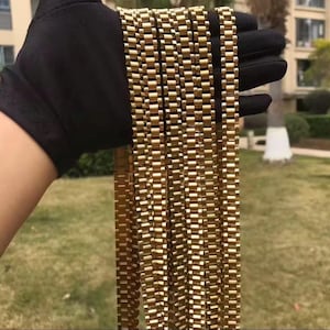 May include: A collection of gold-coloured, rectangular link chains held in a black gloved hand. The chains are displayed against a blurred outdoor background with green grass and trees.
