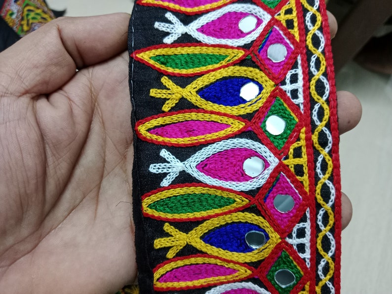May include: Black fabric trim with colorful embroidery and small round mirrors. The trim features a repeating pattern of fish shapes, geometric designs, and a border of small circles.