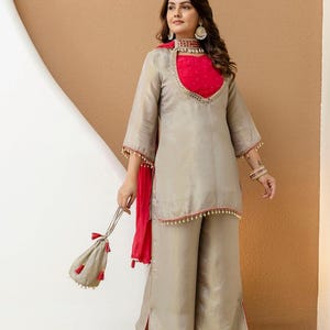 Könnte beinhalten: Beiges Ensemble aus Tunika und weiten Hosen. Die Tunika hat einen roten Ausschnitt und Ärmeldetails. Ein passender roter Schal und eine kleine Tasche mit Quasten vervollständigen das Outfit. Geeignet für besondere Anlässe.