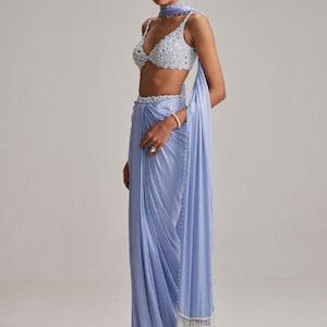 May include: A light blue saree ensemble featuring a beaded crop top, matching draped skirt, and a scarf. The saree has a beaded belt and a fringed edge. The outfit is a soft, flowing fabric, perfect for special occasions.