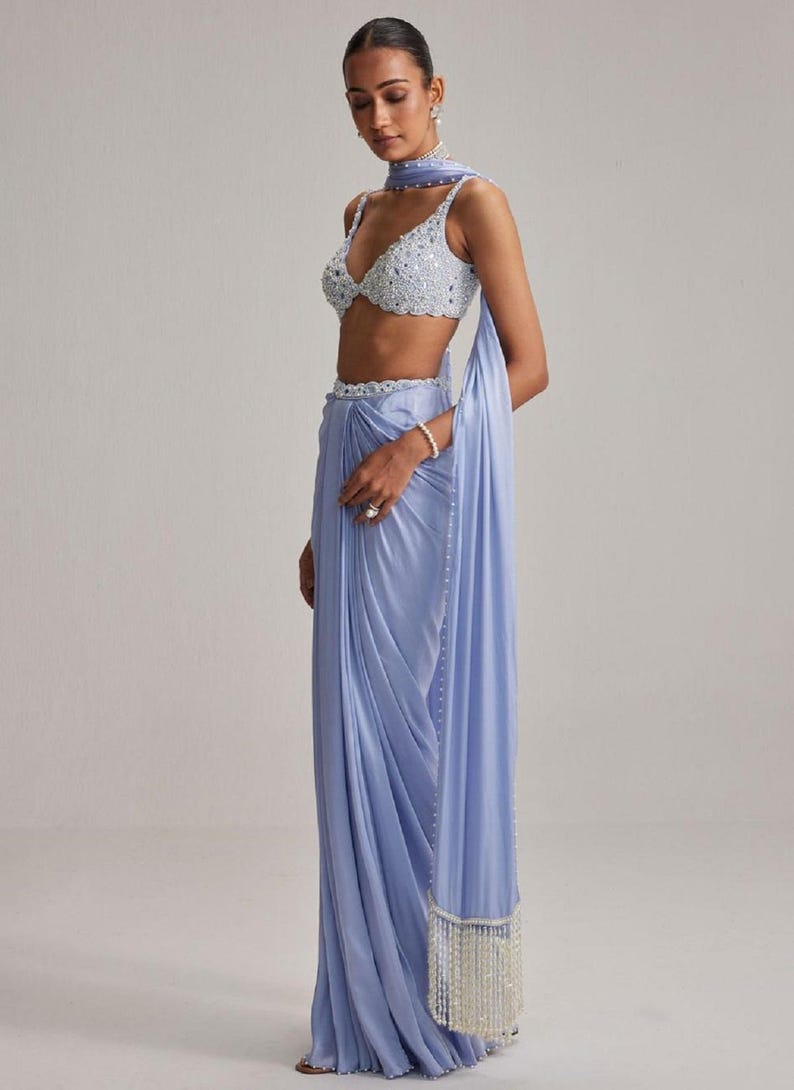 May include: A light blue saree ensemble featuring a beaded crop top, matching draped skirt, and a scarf. The saree has a beaded belt and a fringed edge. The outfit is a soft, flowing fabric, perfect for special occasions.