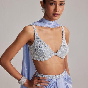 May include: A light blue beaded top and matching draped skirt. The top features a scalloped edge and is adorned with small beads and mirrored accents. A matching belt and scarf complete the ensemble. A silver bracelet and ring are also visible.