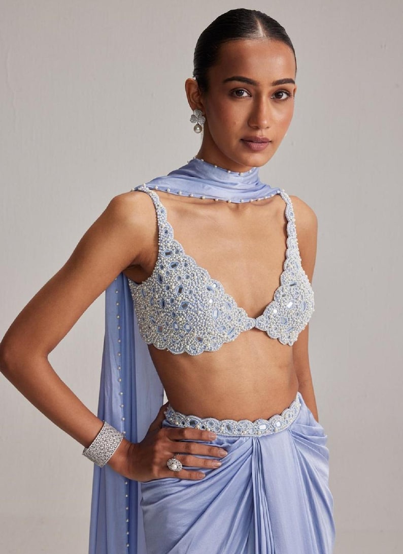 May include: A light blue beaded top and matching draped skirt. The top features a scalloped edge and is adorned with small beads and mirrored accents. A matching belt and scarf complete the ensemble. A silver bracelet and ring are also visible.
