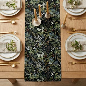 May include: A table setting with a black floral table runner. The runner features a pattern of green, white, and blue leaves and flowers. Gold-colored cutlery and white plates with gold rims are set on a light wood table. Beige napkins and candles complete the elegant look.
