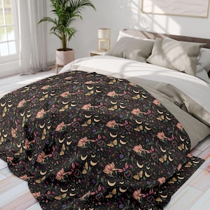 Op de afbeelding: Een zwarte deken met een herhalend patroon van roze bloemen, gele vlinders, halve manen en botanische elementen. De deken ligt op een bed met neutraal gekleurd beddengoed.