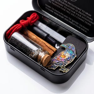 May include: A black metal tin containing a colorful enamel raven pin, a glass vial filled with black sand and a cork stopper, a cinnamon stick, a black candle, and a bundle of red yarn. The lid features text about protection and talismans.