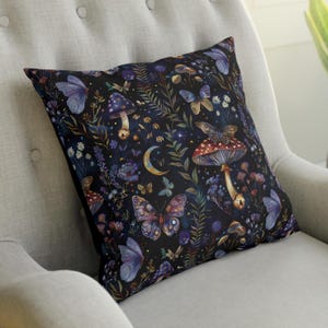 Peut inclure: Coussin carré avec un fond sombre orné d'un motif fantaisiste de papillons, de champignons, de fleurs et d'un croissant de lune. Le coussin a une bordure noire et est posé sur une chaise grise.