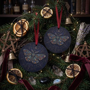 May include: Two circular dark blue ornaments featuring a detailed moth illustration with colorful floral patterns and geometric gold-toned linework. Each ornament hangs from a red ribbon, surrounded by dried orange slices, small brass bells, and crystals.