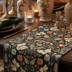 Könnte beinhalten: Ein gedeckter Esstisch mit einem schwarzen Blumen-Tischläufer mit Schmetterlingen und Blumen. Goldene Kerzenständer, Vasen und eine grüne Serviette ergänzen die Dekoration. Angezündete Kerzen sorgen für ein warmes Ambiente.