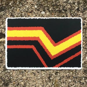 May include: A rectangular sticker with a black background and a design featuring a yellow chevron and red horizontal lines. The sticker has a white border and is placed on a textured, gray surface.