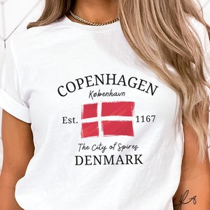 May include: White t-shirt with a graphic design featuring the text "Copenhagen", "Kobenhavn", "Est. 1167", "The City of Spires", and "DENMARK". The graphic also includes a red and white Danish flag.