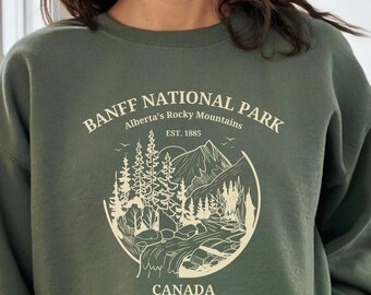 May include: A military green sweatshirt with a cream colored graphic of a mountain scene and the text "Banff National Park, Alberta's Rocky Mountains, Est. 1885, Canada".