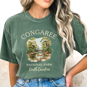 May include: A green t-shirt with a vintage-style illustration of a river scene and the text "Congaree National Park South Carolina".