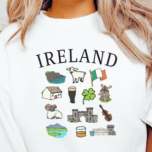 May include: White sweatshirt with the word "Ireland" above a collection of illustrated Irish icons, including a sheep, flag, and landmarks. The design features a green shamrock, a pint of stout, and a violin.