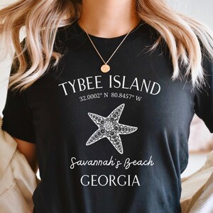 May include: Black t-shirt with white text that reads "Tybee Island 32.0002&deg; N 80.8457&deg; W Savannah's Beach GEORGIA" and a stylized white starfish graphic.