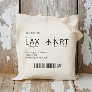 May include: White canvas tote bag with a black printed boarding pass design. The boarding pass shows a flight from LAX Los Angeles to NRT Tokyo Narita. The text also includes the passenger name, class, and boarding time.