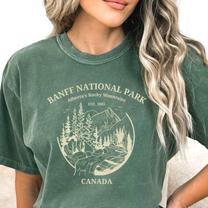 May include: A green t-shirt with a vintage-style graphic of Banff National Park, Alberta's Rocky Mountains. The graphic includes a mountain range, trees, a river, and the text "Banff National Park, Alberta's Rocky Mountains, Est. 1885, Canada".