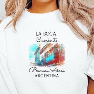 May include: White sweatshirt with a watercolor illustration of colorful buildings in Buenos Aires, Argentina. The text "LA BOCA Caminito Buenos Aires ARGENTINA" is printed on the shirt.