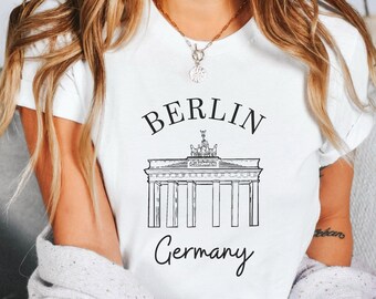 May include: White t-shirt featuring a black and white illustration of the Brandenburg Gate in Berlin, Germany. The word "BERLIN" is above the gate, and "Germany" is below it, in a handwritten font. The shirt is a crew neck.