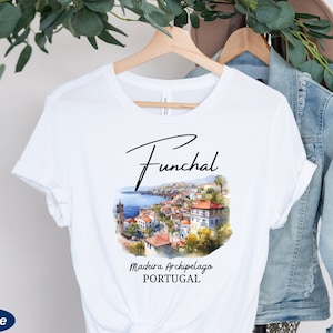 May include: White t-shirt with a black and white illustration of the city of Funchal, Madeira Archipelago, Portugal. The text "Funchal" is written in a cursive font above the illustration. The text "Madeira Archipelago" and "PORTUGAL" are written below the illustration.