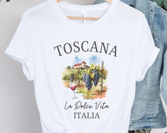 May include: White t-shirt with a graphic of a vineyard and a glass of wine. The text "Toscana La Dolce Vita Italia" is printed on the shirt.